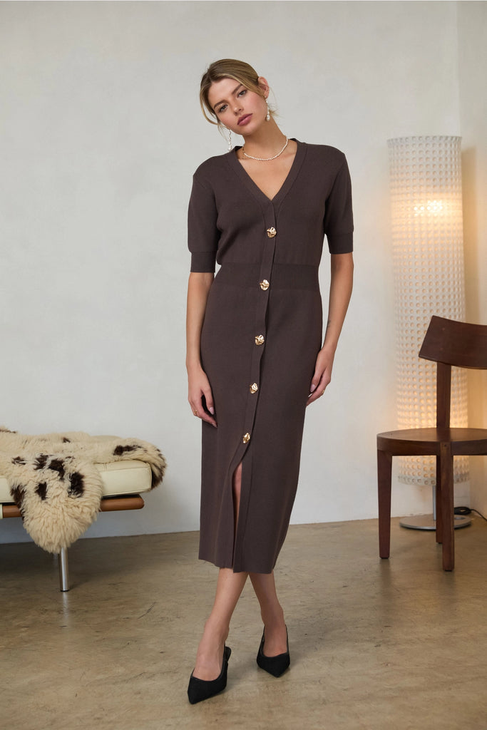 Woman wearing a long brown dress with short sleeves, a V-Neck cut, and gold buttons in doors