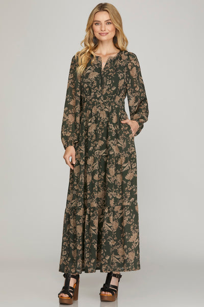 Woman wearing a long earthy green floral maxi dress with pockets on a plain background