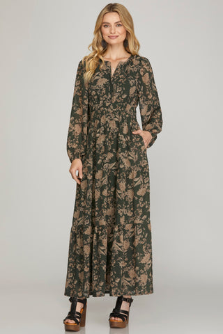 Woman wearing a long earthy green floral maxi dress with pockets on a plain background