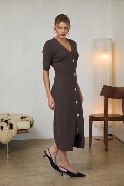 Woman wearing a long brown dress with short sleeves, a V-Neck cut, and gold buttons in doors