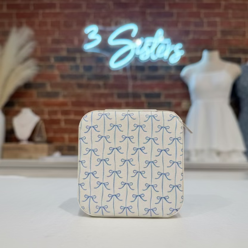 Compact jewelry box with blue bow pattern on a white surface, with '3 Sisters' neon sign in the background.