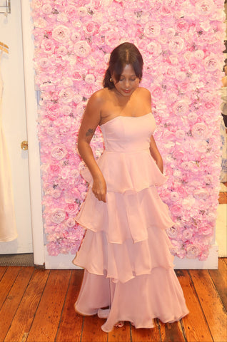 Woman in a pink dress standing against a pink flower backdrop with a wooden floor.