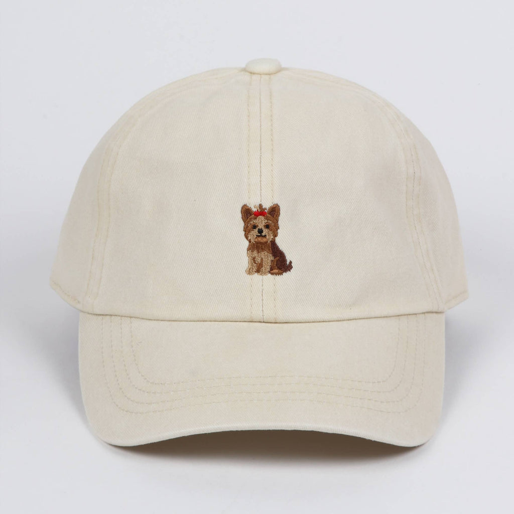 Off-white cotton baseball cap featuring a small embroidered Yorkshire terrier dog wearing a red bow on the front center, against a plain background