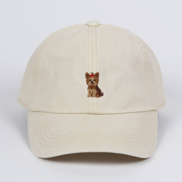 Off-white cotton baseball cap featuring a small embroidered Yorkshire terrier dog wearing a red bow on the front center, against a plain background