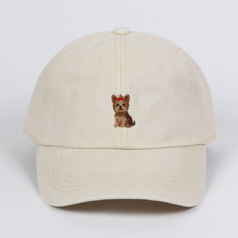 Off-white cotton baseball cap featuring a small embroidered Yorkshire terrier dog wearing a red bow on the front center, against a plain background