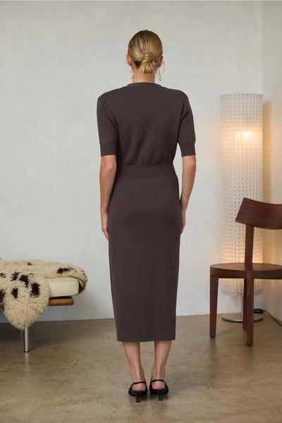 Woman wearing a long brown dress with short sleeves, a V-Neck cut, and gold buttons in doors