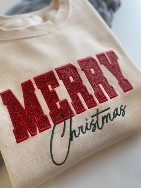 A cream-colored crewneck sweatshirt featuring 'MERRY Christmas' printed on the front, with 'MERRY' in glittery red block letters and 'Christmas' in dark green cursive script