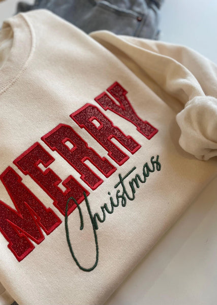 A cream-colored crewneck sweatshirt featuring 'MERRY Christmas' printed on the front, with 'MERRY' in glittery red block letters and 'Christmas' in dark green cursive script