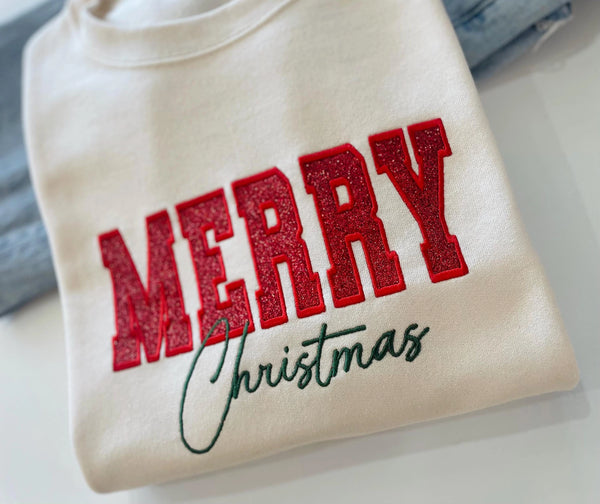 A cream-colored crewneck sweatshirt featuring 'MERRY Christmas' printed on the front, with 'MERRY' in glittery red block letters and 'Christmas' in dark green cursive script