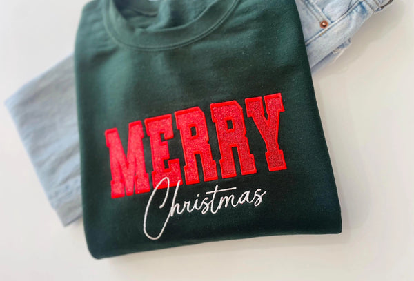 A dark green-colored crewneck sweatshirt featuring 'MERRY Christmas' printed on the front, with 'MERRY' in glittery red block letters and 'Christmas' in white cursive script