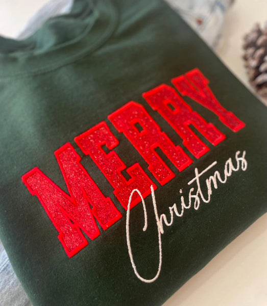 A dark green-colored crewneck sweatshirt featuring 'MERRY Christmas' printed on the front, with 'MERRY' in glittery red block letters and 'Christmas' in white cursive script