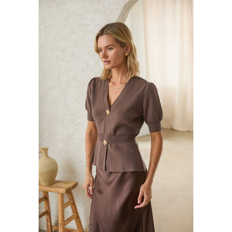 Woman wearing a brown short-sleeve top with gold buttons and a brown skirt indoors.