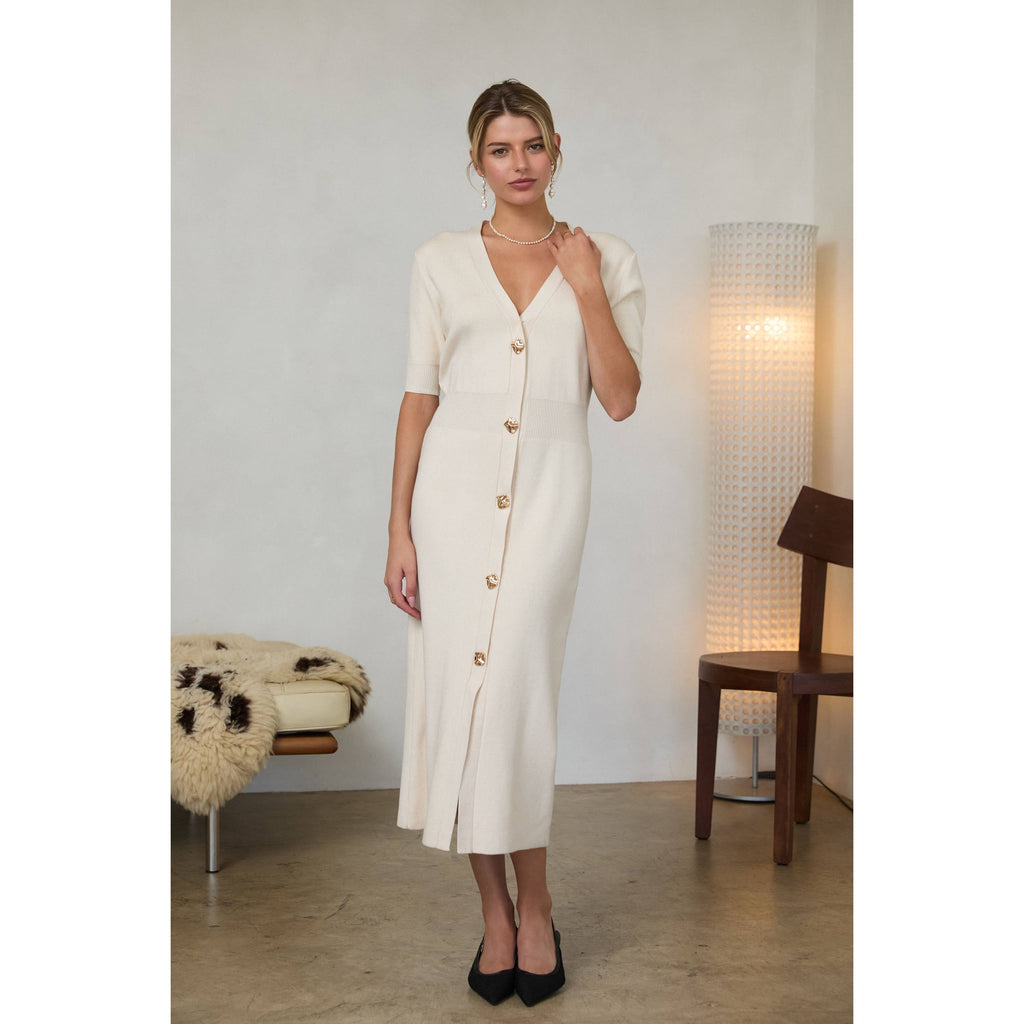 Woman wearing a cream-colored midi dress with short puff sleeves, a V-neckline, and a front button placket with gold buttons, standing indoors.