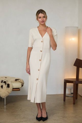 Woman wearing a cream-colored midi dress with short puff sleeves, a V-neckline, and a front button placket with gold buttons, standing indoors.