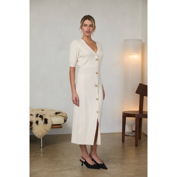 Woman wearing a cream-colored midi dress with short puff sleeves, a V-neckline, and a front button placket with gold buttons, standing indoors.