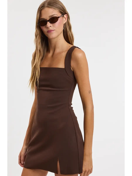 A woman modeling a sleeveless brown mini dress with a square neckline and a side slit, paired with dark sunglasses, against a plain light gray background.