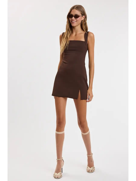 A woman modeling a sleeveless brown mini dress with a square neckline and a side slit, paired with dark sunglasses, against a plain light gray background.