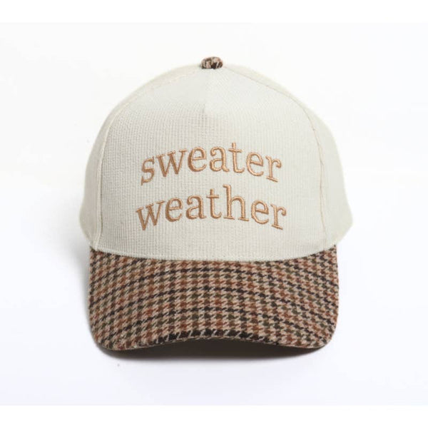 A baseball cap with an off-white crown and a brown, beige, and black houndstooth patterned visor. The front of the cap features the embroidered text 'sweater weather' in a tan text
