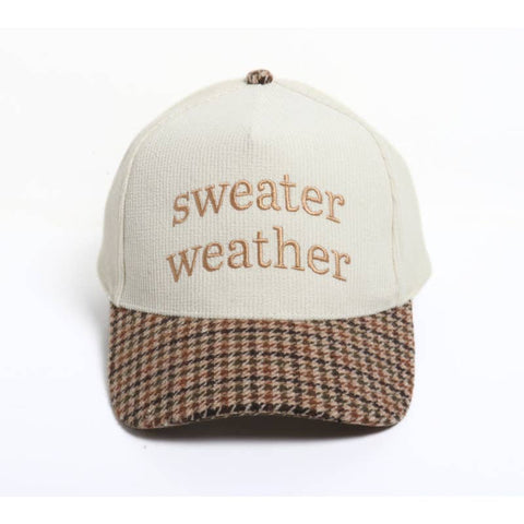 A baseball cap with an off-white crown and a brown, beige, and black houndstooth patterned visor. The front of the cap features the embroidered text 'sweater weather' in a tan text